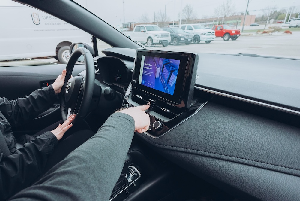 A person sits in the driver's seat of a car, holding the steering wheel with one hand and using the touchscreen display on the center console with the other hand. Several parked vehicles are visible outside.