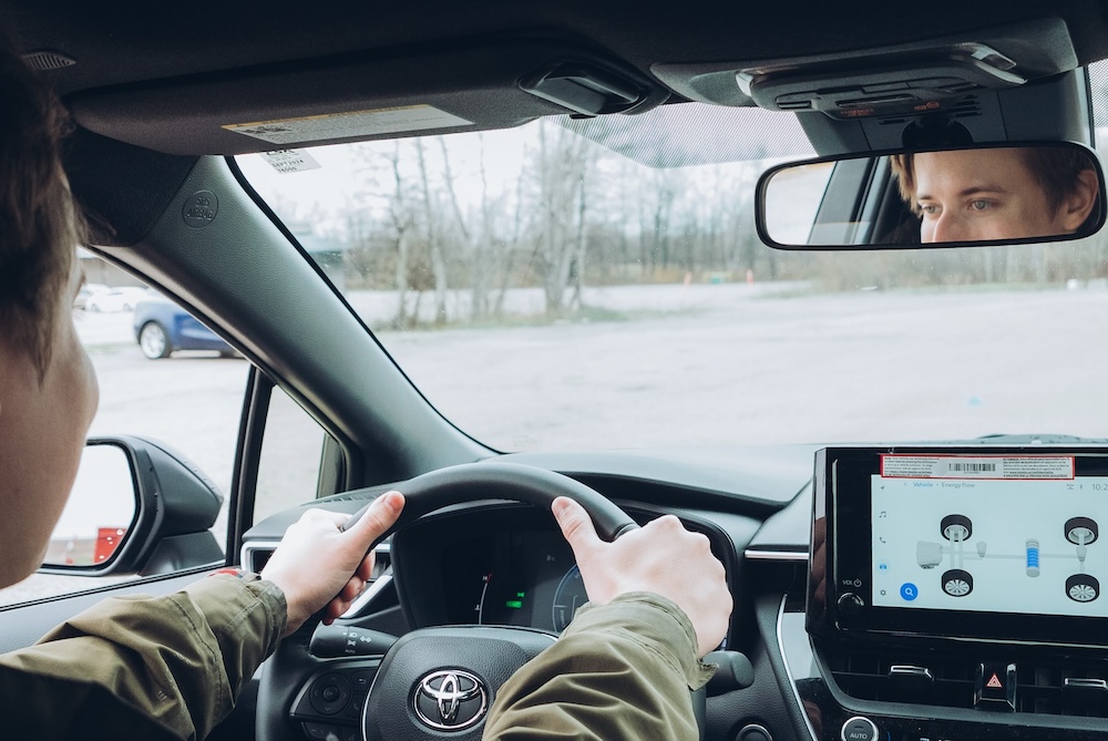 A person sits in a driving simulator with three large screens showing a virtual urban street scene with vehicles, pedestrians, and buildings. The person holds a steering wheel and looks at the central screen.