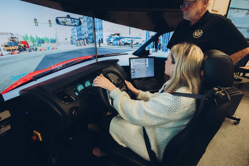 Three adults sit at desks in a classroom, holding remote controls and facing forward. Papers, notebooks, and name cards are on the desks. A computer monitor and office supplies are visible in the background.