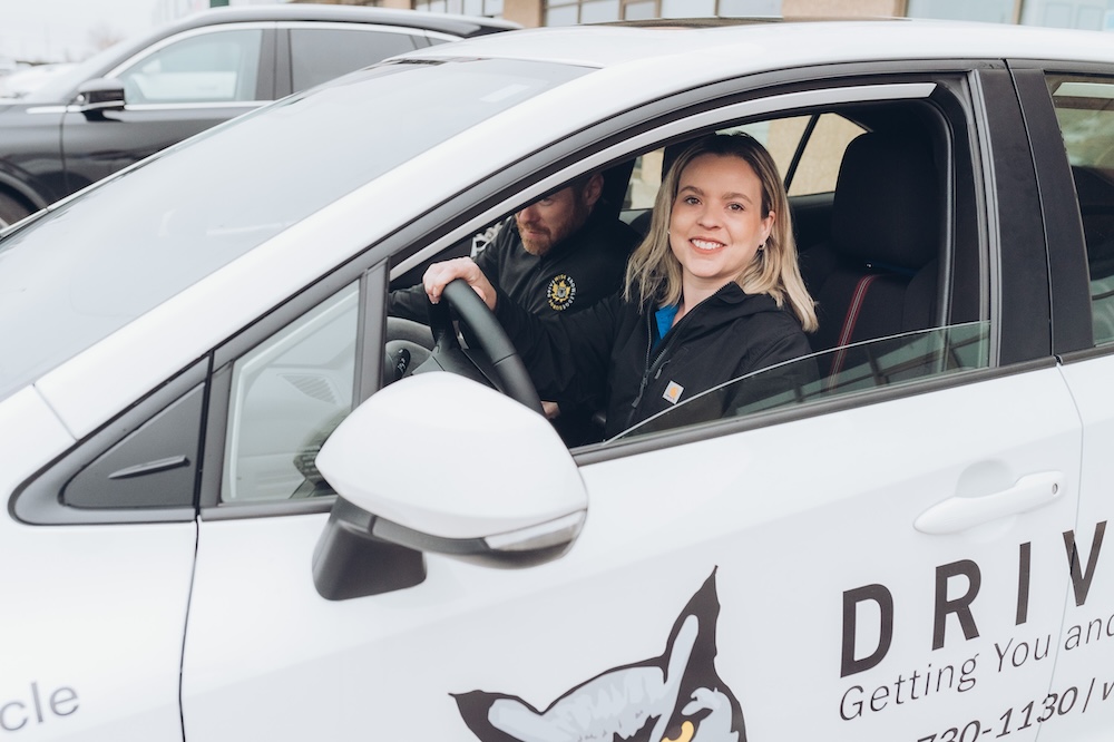 A person is driving a car while another person in the passenger seat holds a clipboard with a form, writing with a green pen. The scene appears to be a driving test or evaluation.