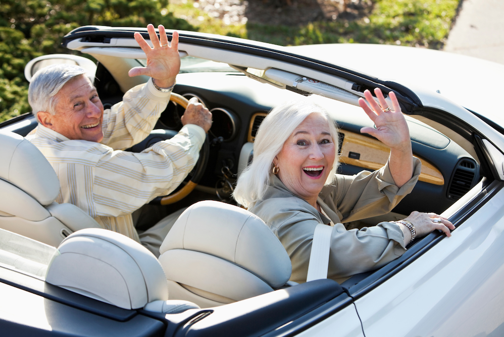 Three adults sit at desks in a classroom, holding remote controls and facing forward. Papers, notebooks, and name cards are on the desks. A computer monitor and office supplies are visible in the background.