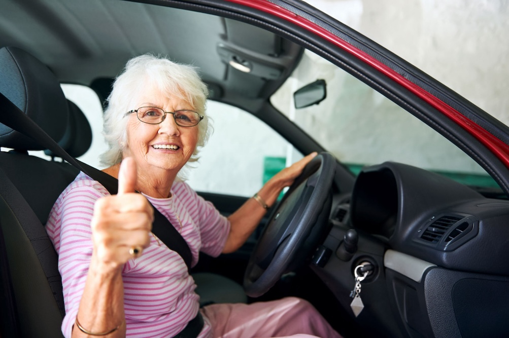 A person sits in a driving simulator with three large screens showing a virtual urban street scene with vehicles, pedestrians, and buildings. The person holds a steering wheel and looks at the central screen.