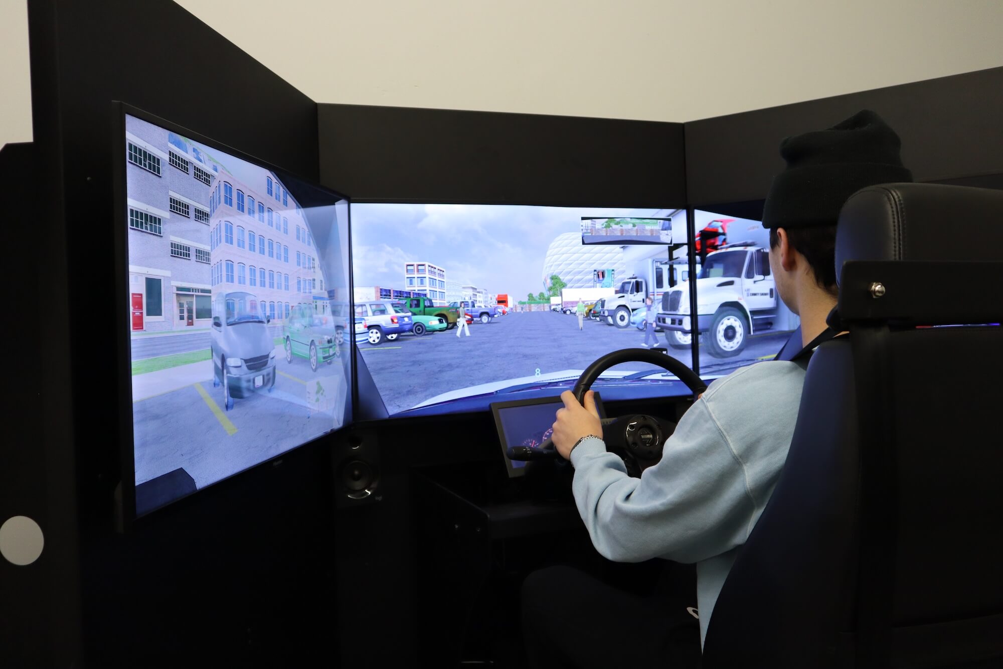 A person sits in a driving simulator with three large screens showing a virtual urban street scene with vehicles, pedestrians, and buildings. The person holds a steering wheel and looks at the central screen.