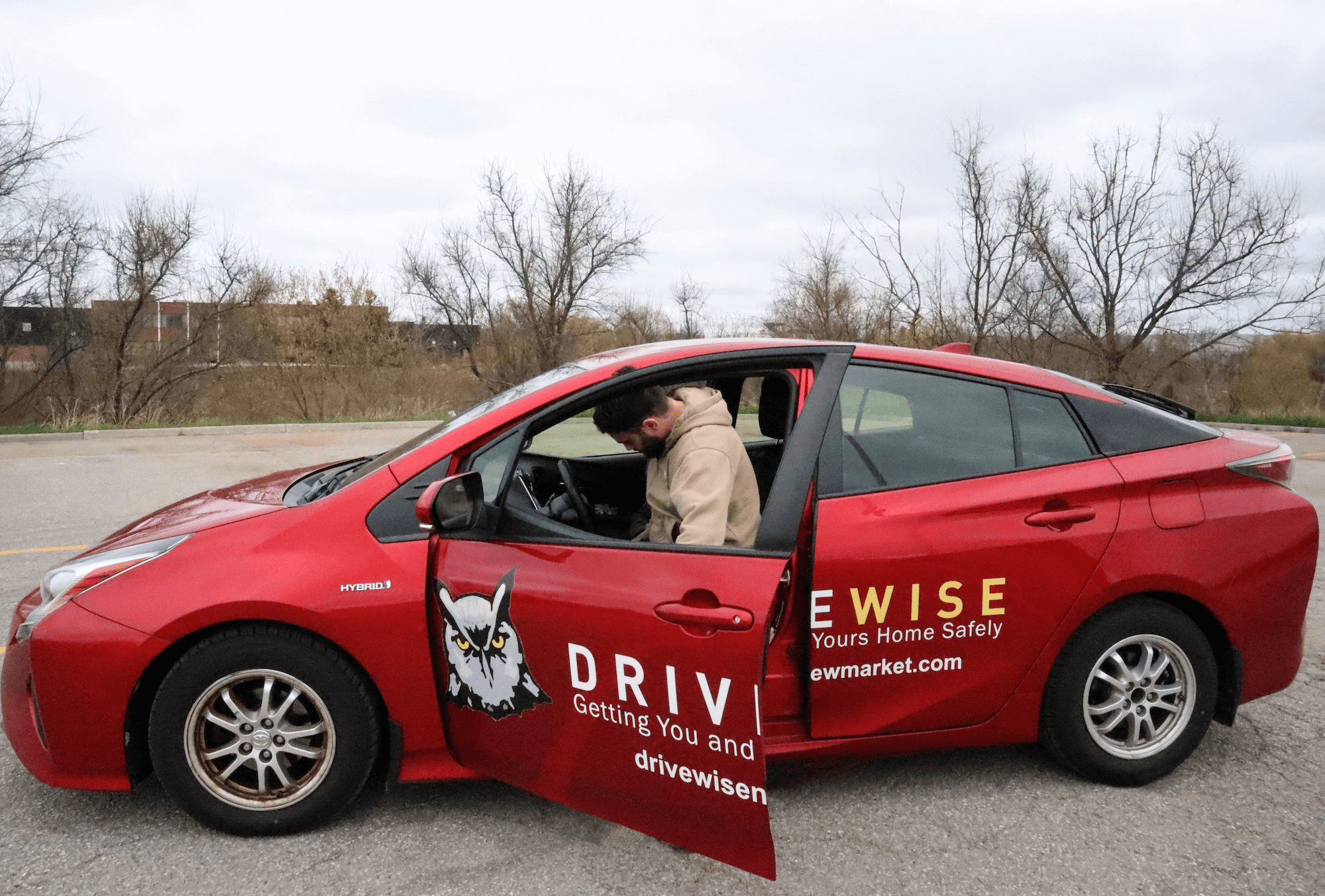 A person sits in the driver's seat of a red sedan parked outdoors. The car has "DRIVEWISE" branding and an owl graphic on the side, along with the slogan "Getting You and Your Home Safely.