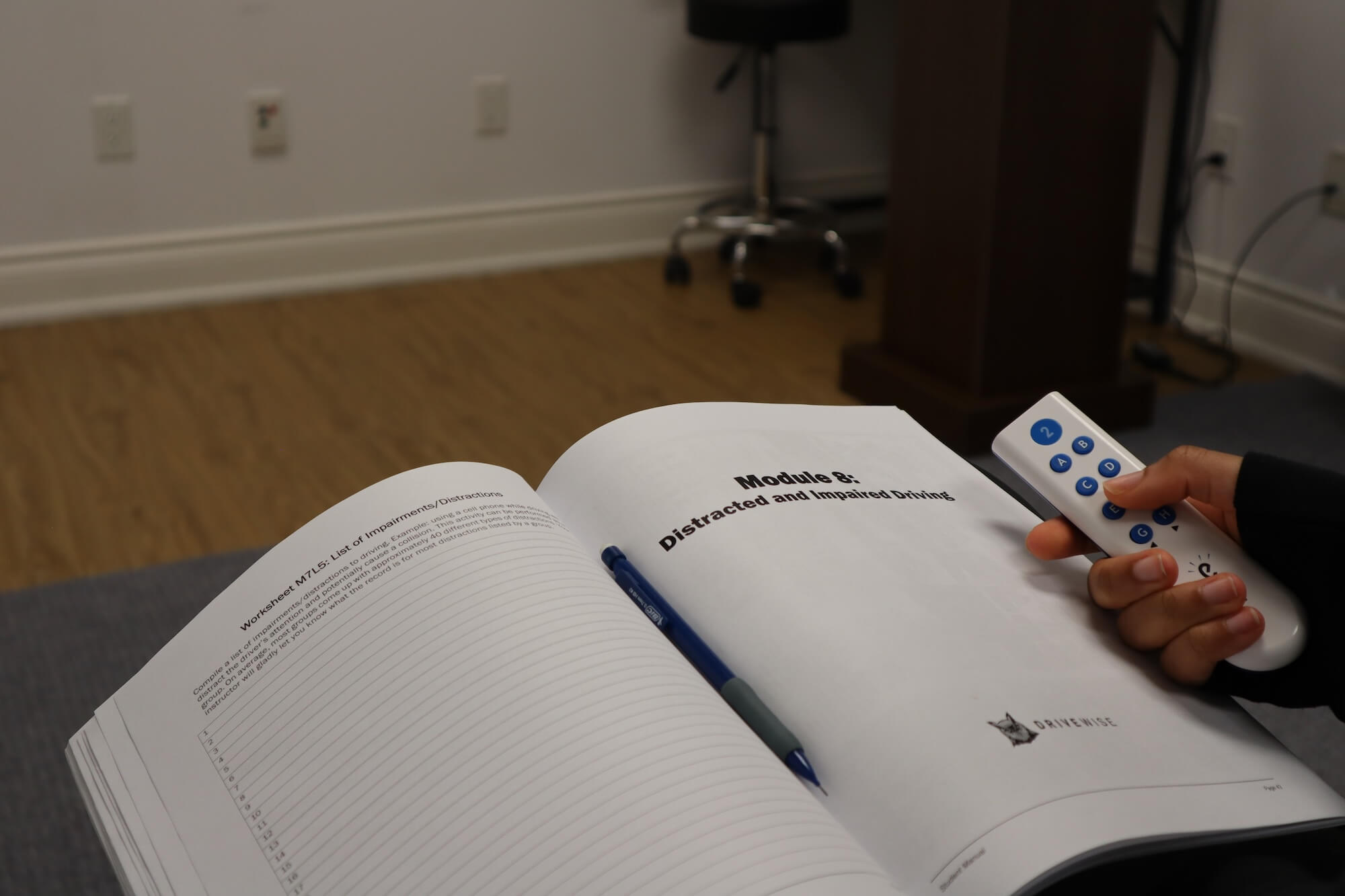 A person holds a remote control in their right hand while looking at an open workbook titled "Module 8: Distracted and Impaired Driving" with a pen resting in the book on a wooden desk.