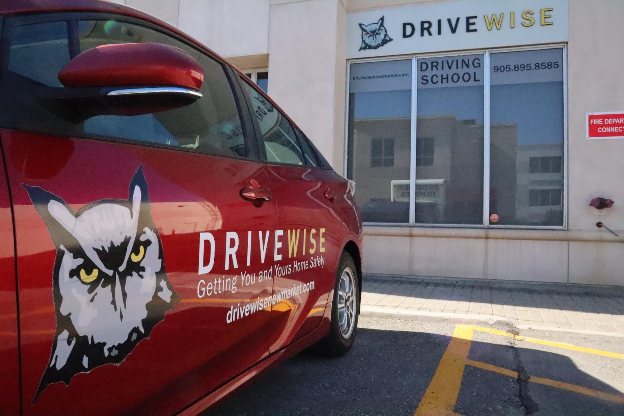 A red car with a DriveWise logo featuring an owl is parked in front of a building with a DriveWise Driving School sign, a phone number, and a red fire department connection notice on the wall.