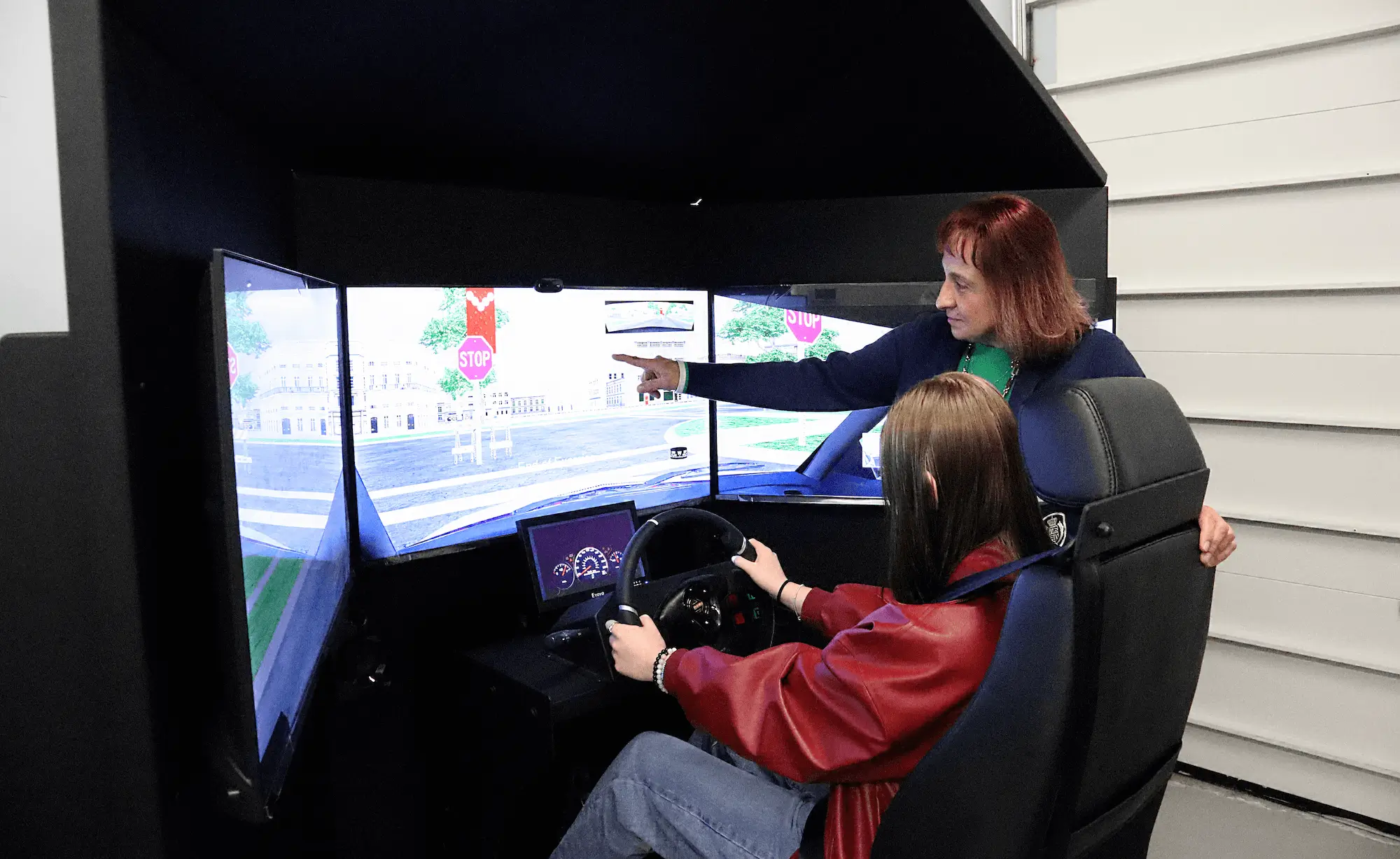 A woman stands beside a seated person using a driving simulator. The simulator has three monitors displaying a virtual road scene. The standing woman points at the screens, appearing to instruct the driver.