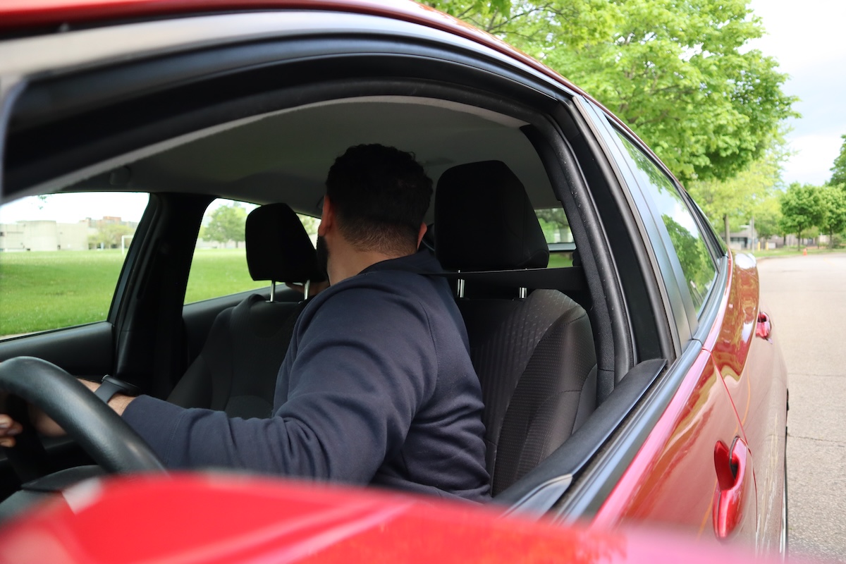A person in a dark hoodie sits in the driver's seat of a red car, looking backward over their shoulder, likely preparing to reverse. The car is parked on a street next to a grassy area with trees.