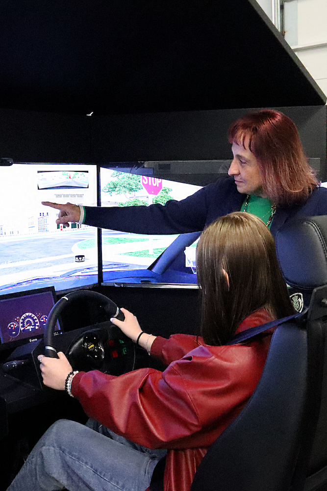 An instructor stands beside a seated person holding a steering wheel in a driving simulator. The instructor points at the simulation screen displaying a street scene and traffic signs.