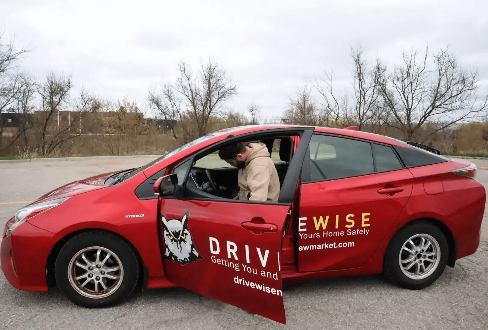 A person in a beige hoodie sits in the driver's seat of a red car with "DRIVEWISE" and an owl logo on the side, parked on a paved surface near leafless trees under a cloudy sky.