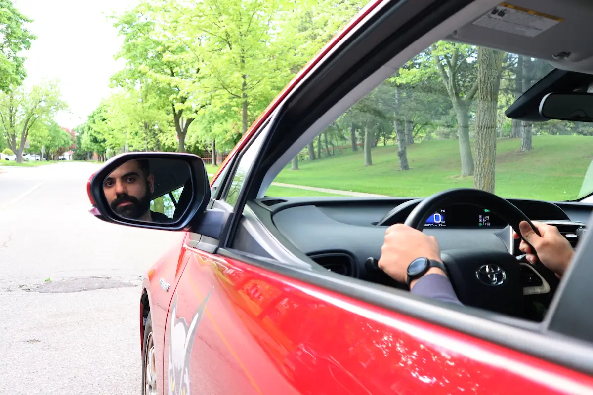 A person sits inside a red car, holding the steering wheel, with their reflection visible in the driver’s side mirror. The car is parked on a tree-lined street with green grass and foliage outside.