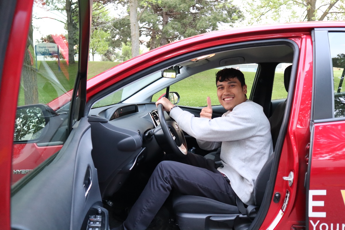 A person sits in the driver’s seat of a red car with the door open, holding the steering wheel and giving a thumbs-up while smiling. There are trees and green grass visible outside the car.