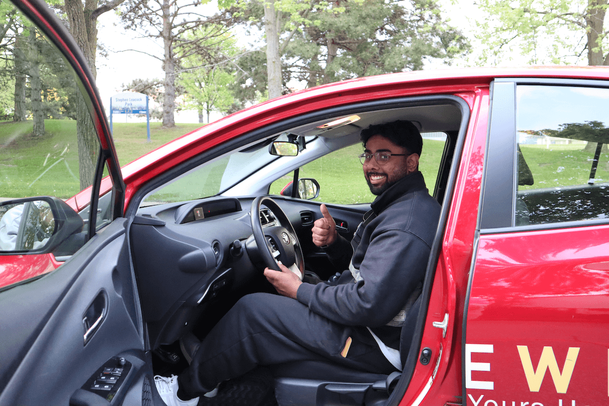 A man sits in the driver's seat of a red car with the door open, smiling and giving a thumbs-up. Trees and grass are visible outside the car.