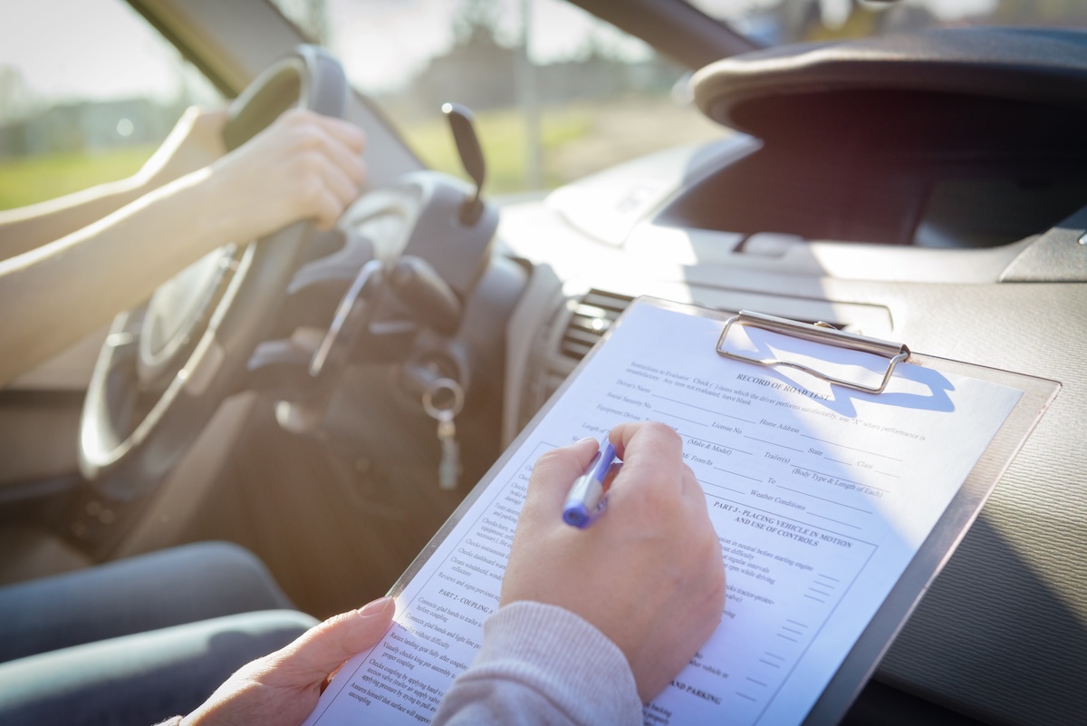 A person holding a clipboard with a form while another person sits in the driver's seat of a car, hands on the steering wheel, during what appears to be a driving test.