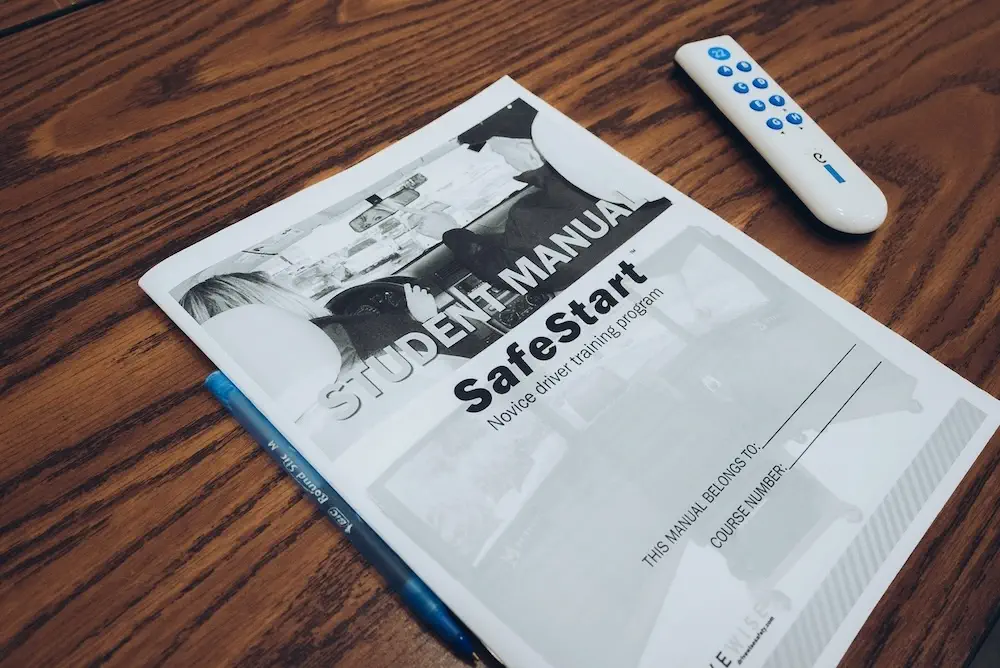 A student manual titled "SafeStart Novice driver training program" lies on a wooden table next to a remote control and a blue pen. The manual cover features black and white images of people in a car.