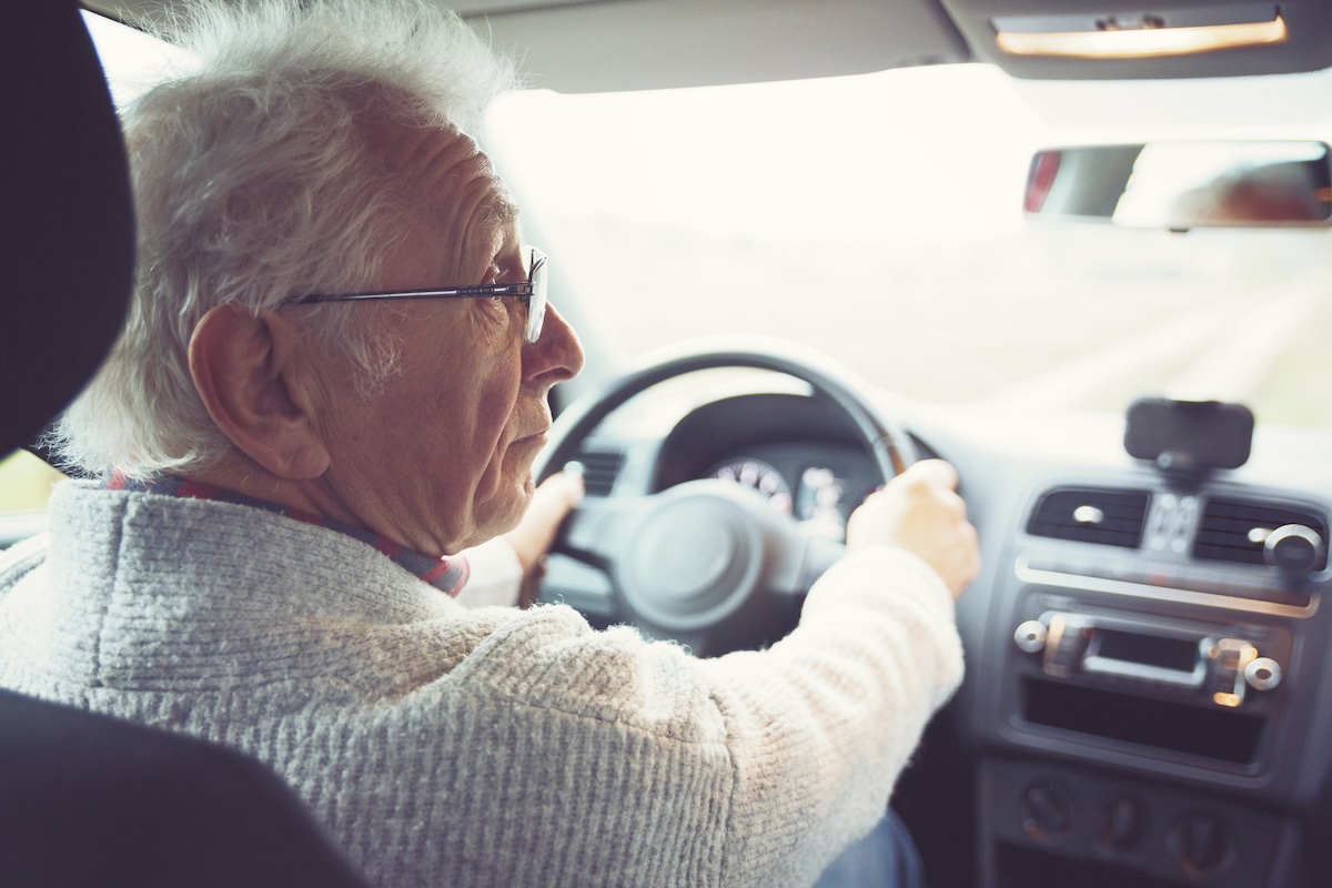 An older man with gray hair, wearing glasses and a sweater, sits in the driver's seat of a car, looking over his shoulder while holding the steering wheel with both hands.